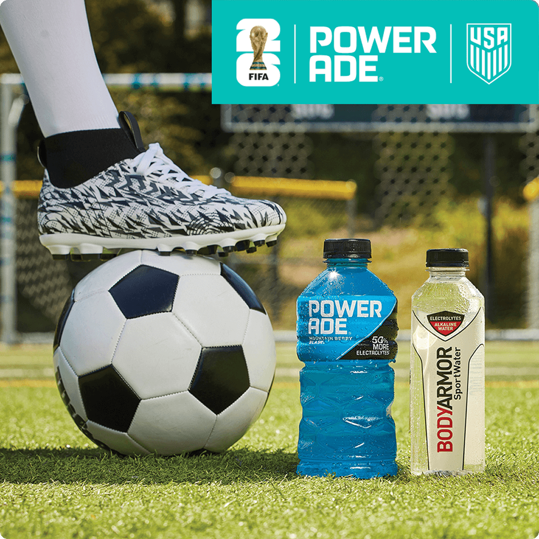 A soccer ball being held by a cleat on a field next to a bottle of Powerade Mountain Berry Blast and a bottle of Bodyarmor